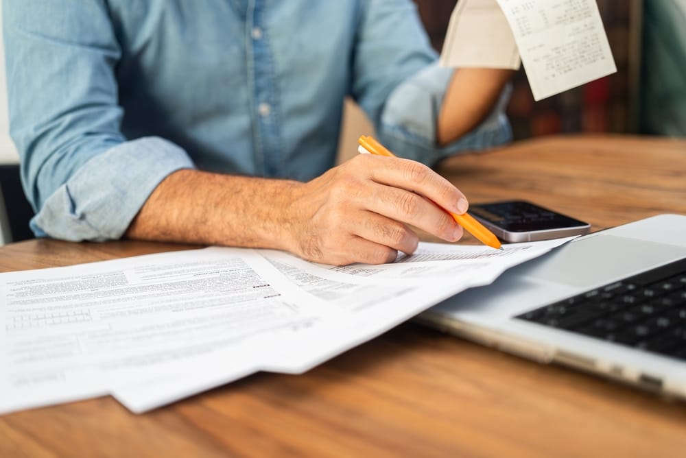 Man in a denim shirt calculating taxes with receipts and forms on a wooden desk. Tax preparation and financial management concept.