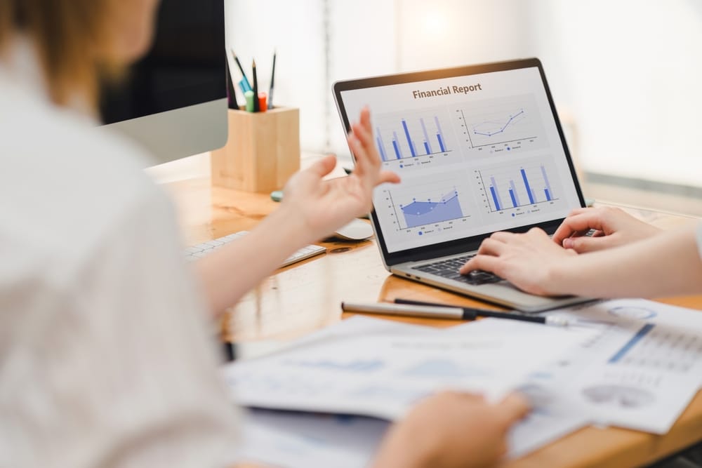 Two businesspeople or an accountant team are analyzing data charts, graphs, and a dashboard on a laptop screen in order to prepare a statistical report and discuss financial data in an office
